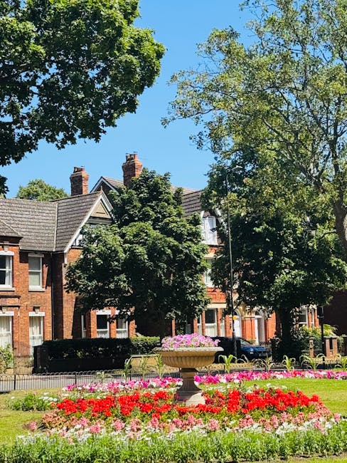 A landscaped urban garden scene featuring a well-maintained grassy area with neatly trimmed lawns and vibrant flower beds containing red, pink, and purple blooms. A large, decorative stone planter fil