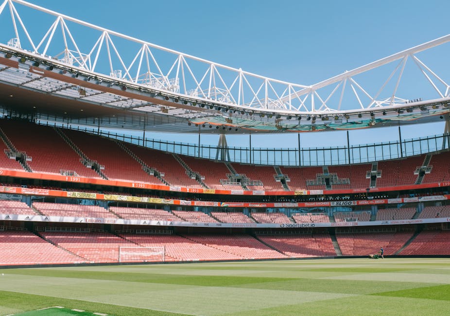 The image shows a large, modern stadium with a well-maintained grass pitch in the foreground, featuring neatly mown, contrasting stripes of green. Surrounding the pitch are multiple tiers of red and p