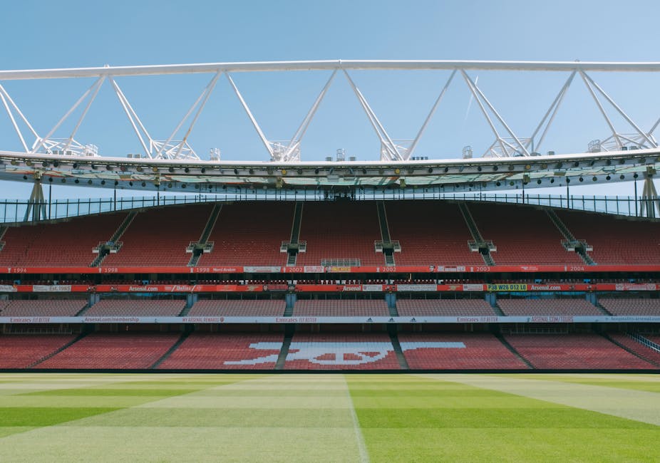 The image shows a large, modern stadium with a well-maintained grass pitch in the foreground, featuring neatly mown, contrasting stripes of green. Surrounding the pitch are multiple tiers of red and p